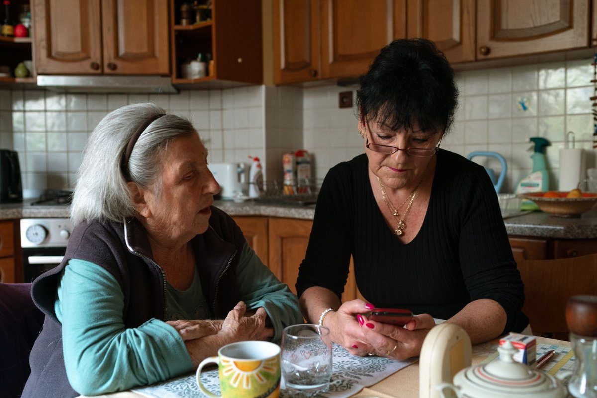A caregiver looks at her phone at a kitchen table while the older woman she visits sits beside her with a cup of tea.
