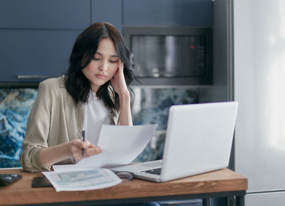 A woman reviews a printed document next to an open laptop at a small office desk.