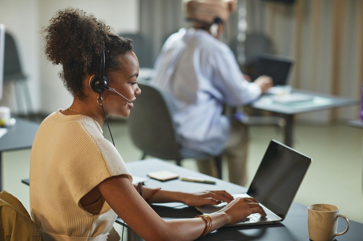 An office staff member wearing a headset types on a laptop at a shared desk while a colleague works in the background.