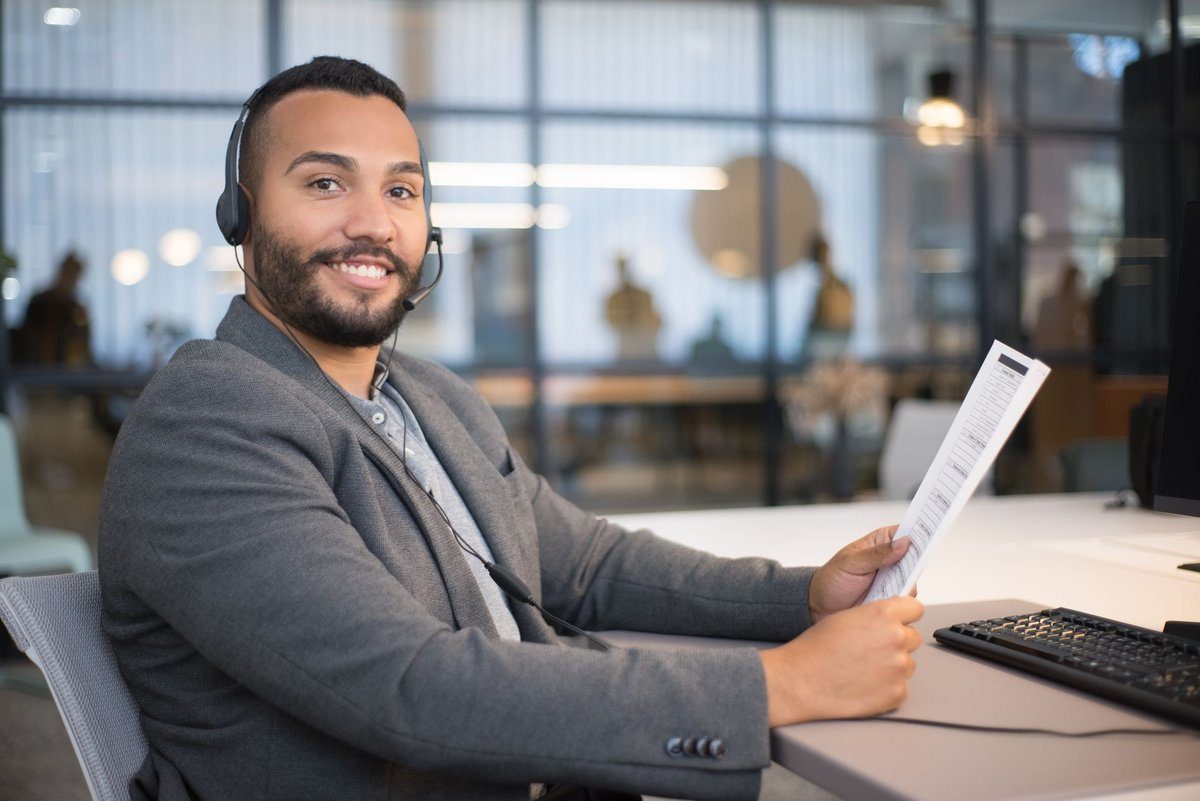A smiling office worker in a headset sits at a desk holding a printed sheet, ready to make calls.