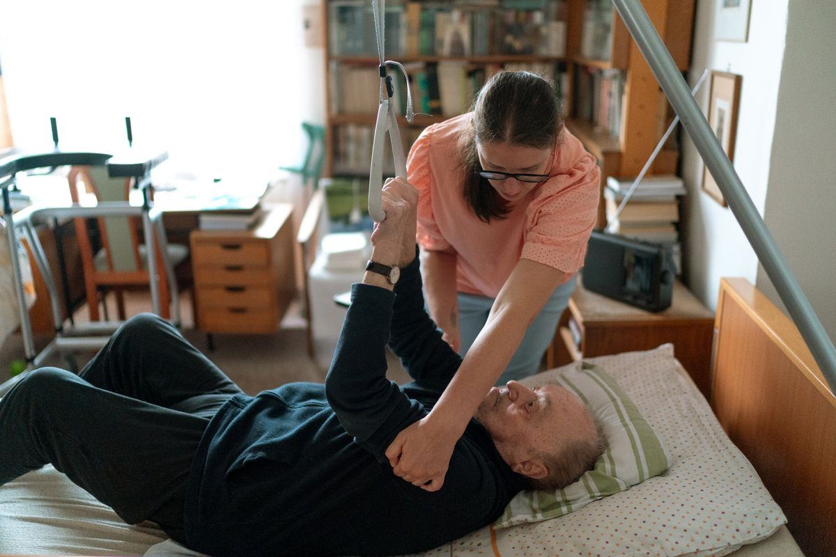 A caregiver in a peach blouse helps an older man lift himself using a trapeze bar over his bed in his book-lined bedroom.