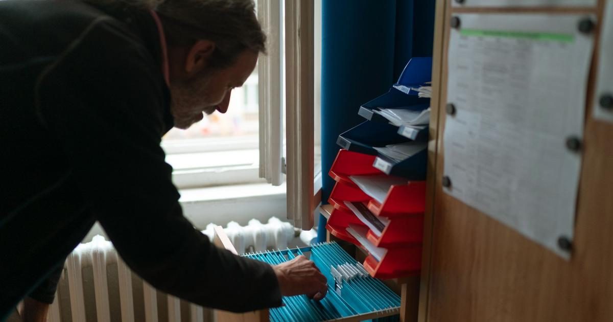 A man leans over an open drawer of blue hanging file folders in a small office, searching through paper records.