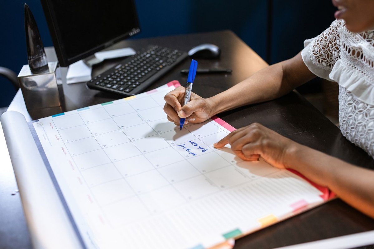 An office worker writes a reminder on a large paper desk calendar next to a computer keyboard.