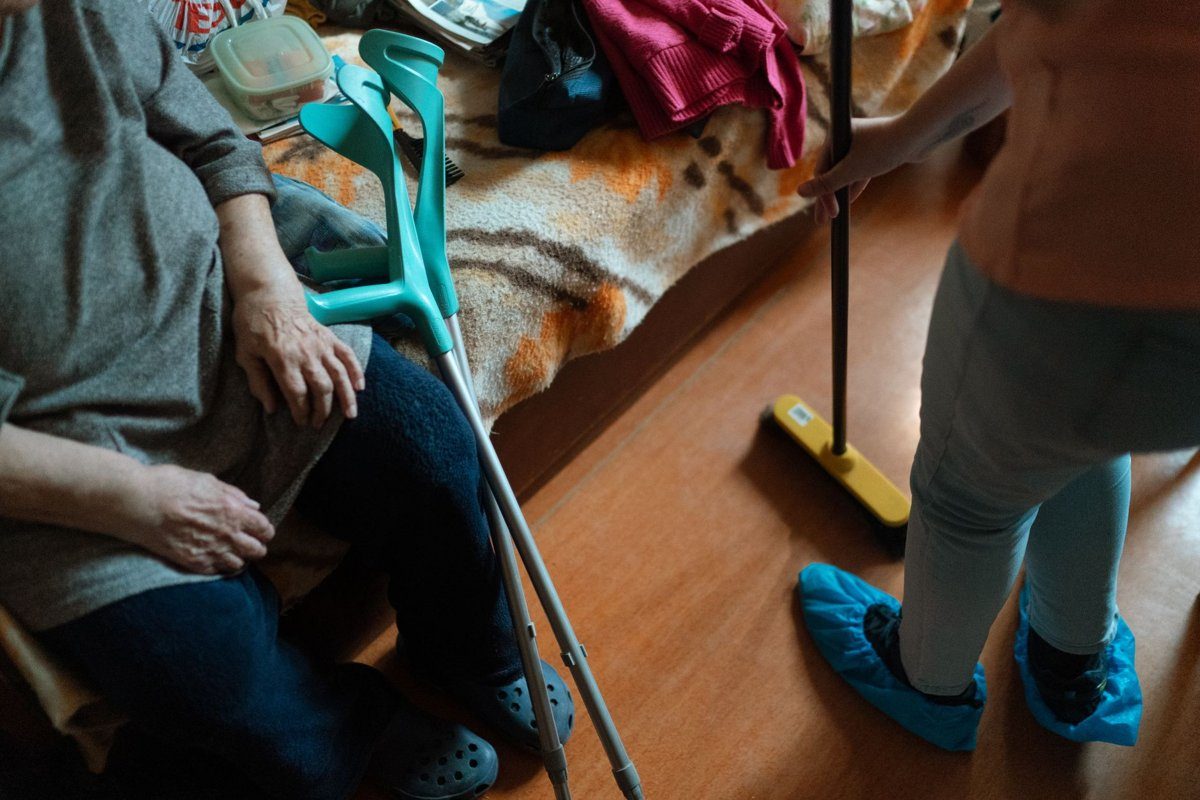 A caregiver sweeps the floor of a bedroom while an older woman with forearm crutches sits on the edge of her bed.