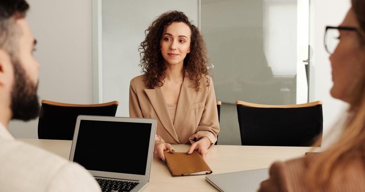 A job candidate in a tan blazer sits across a meeting table from two interviewers with a laptop and a folder in front of her.