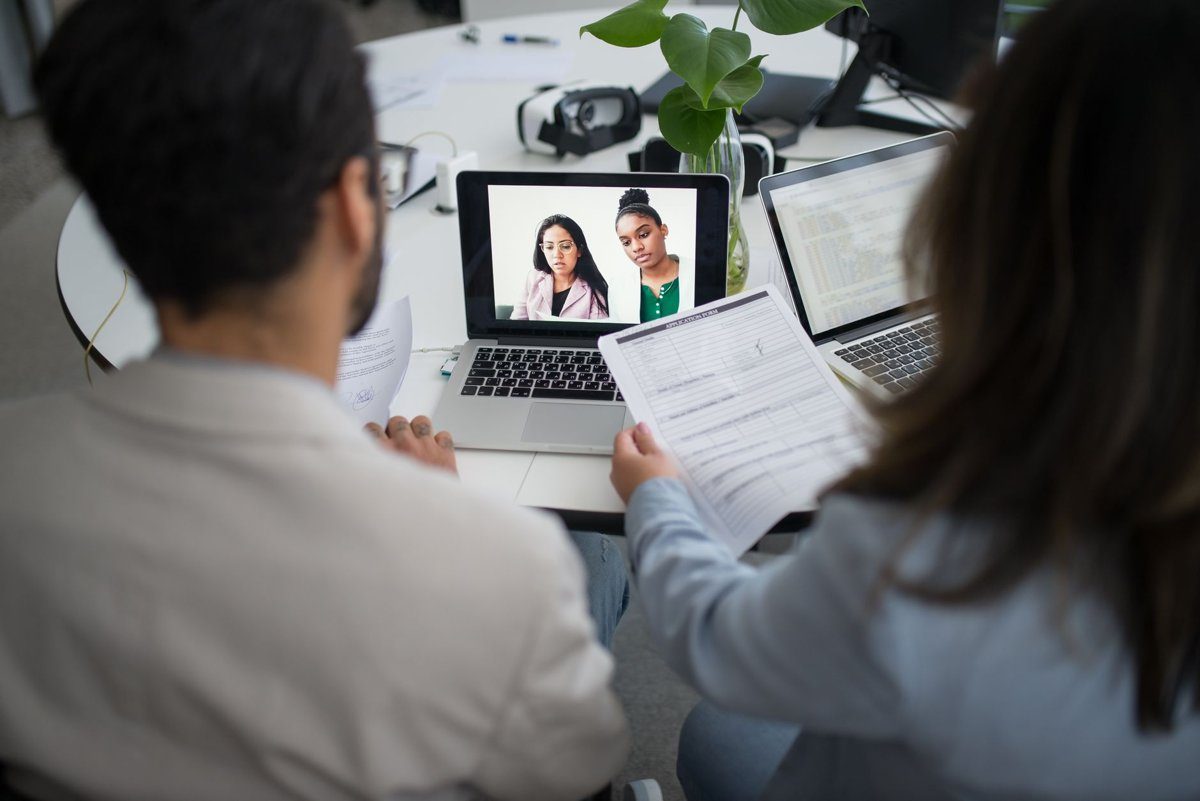 Two hiring staff hold a printed application form while interviewing two candidates on a laptop video call.