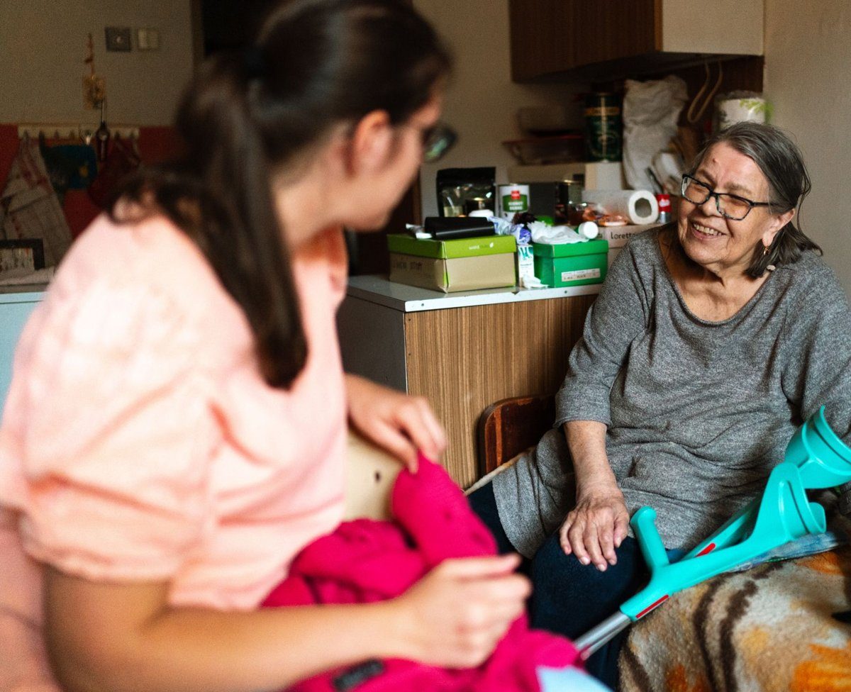 A young caregiver in a pink top folds a red sweater while an older woman with forearm crutches smiles at her from the edge of the bed.