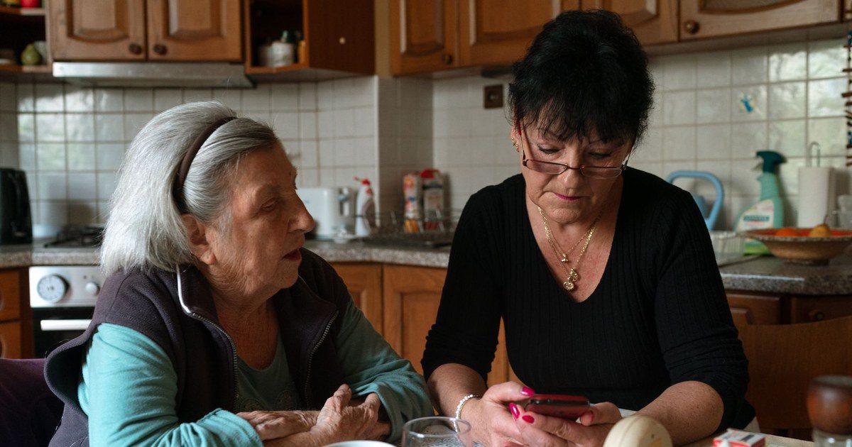 A home care aide in a black sweater sits at a kitchen table with an older woman, checking a phone together over cups of tea.