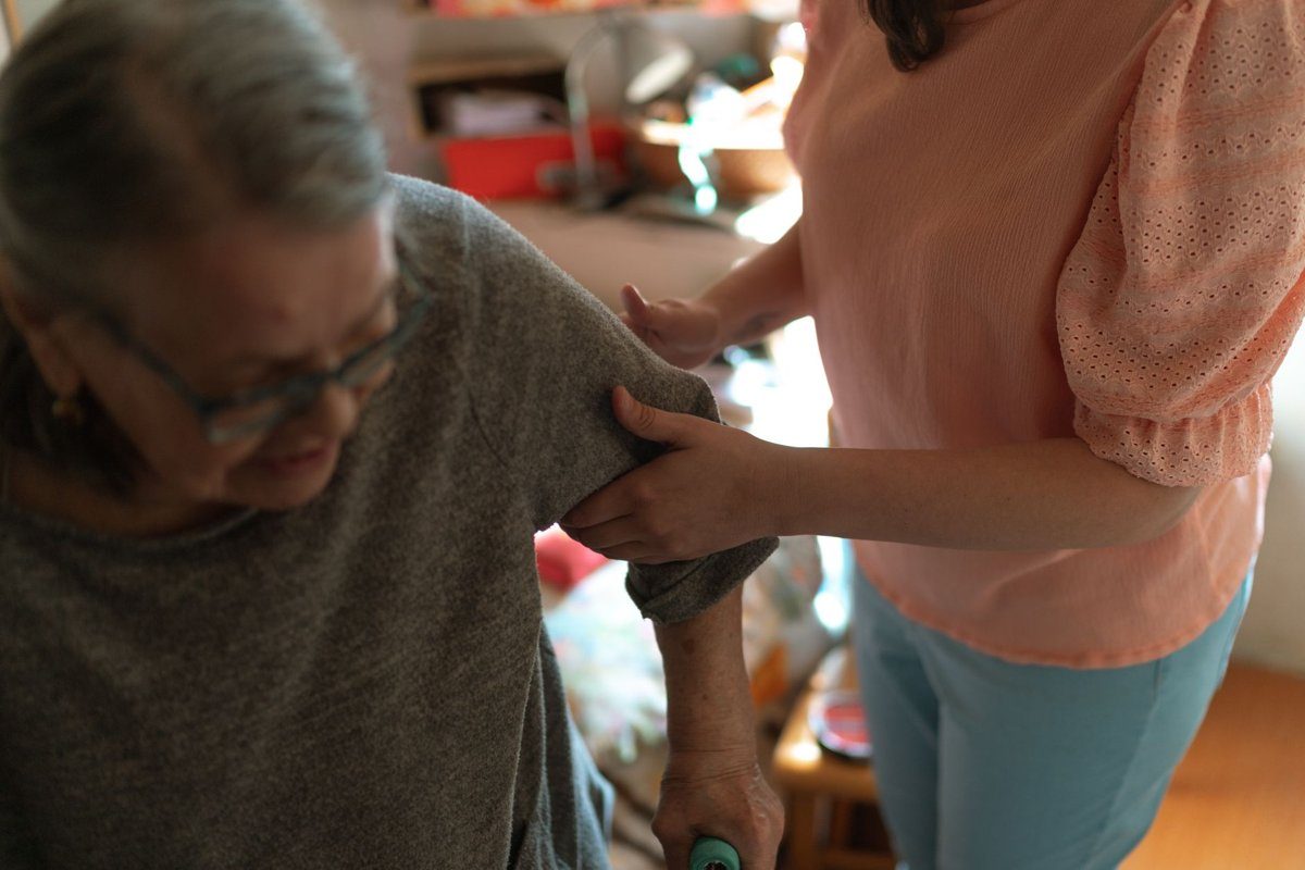 A caregiver steadies an older woman by the arm as she stands up with a walking aid in her home.