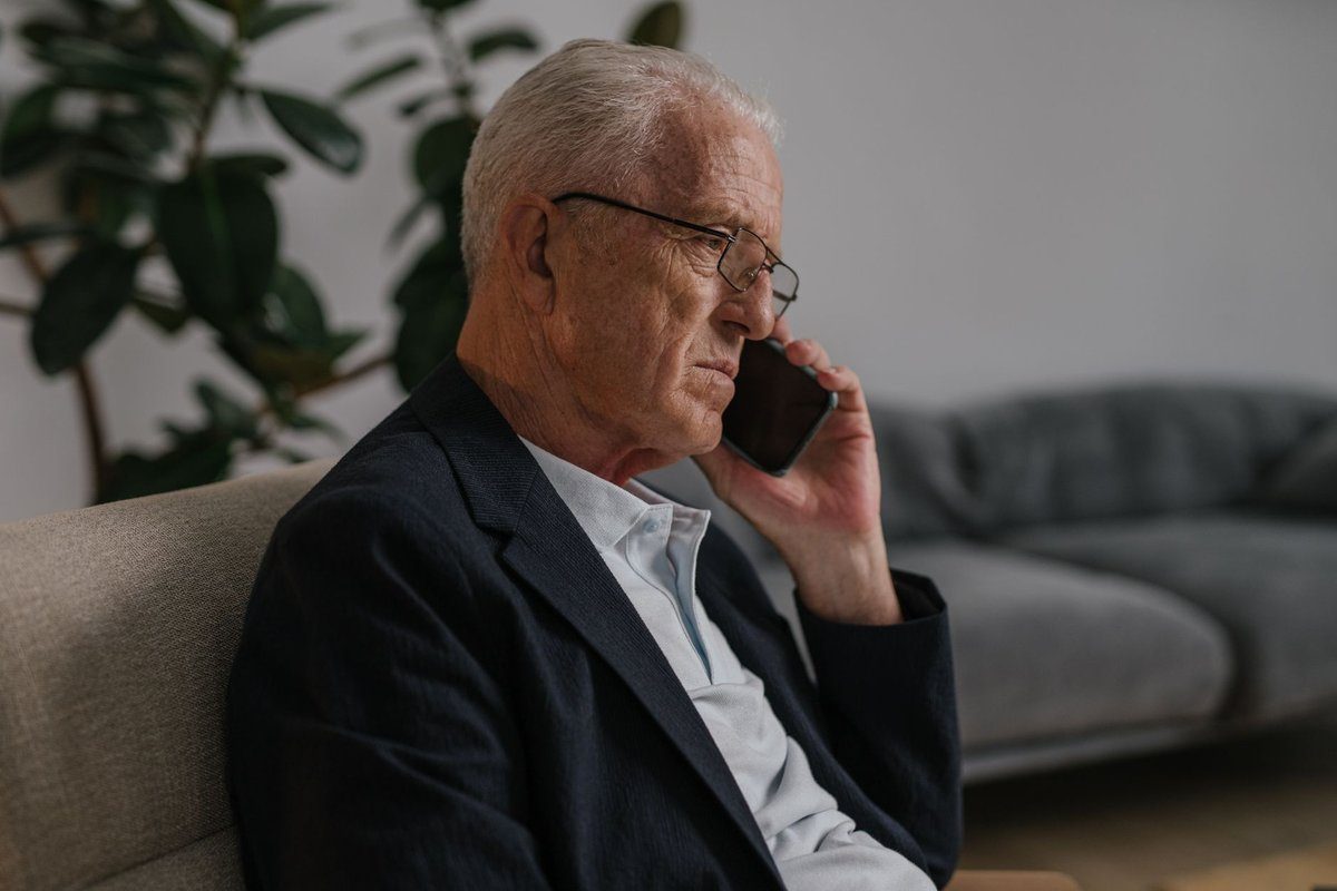 An older man in a dark blazer sits on a sofa at home listening carefully during a phone call.