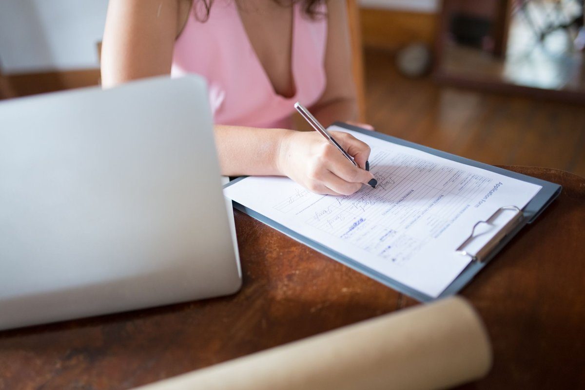 A woman in a pink top writes on a paper form clipped to a clipboard next to a laptop on a wooden table.